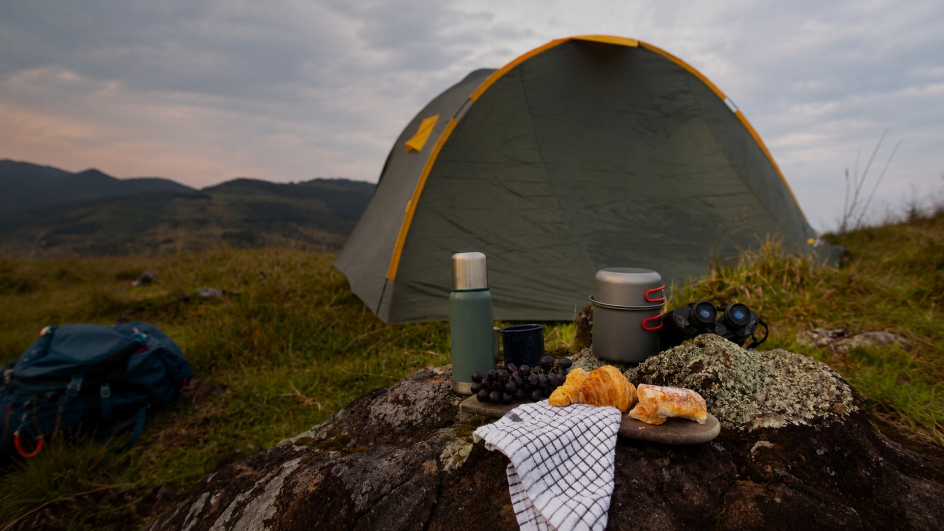 A dome camping tent is pitched on a grassy mountain slope under an overcast sky, with a picnic setup on a rock in the foreground including a thermos, metal pot, bread, grapes, and a checkered towel.