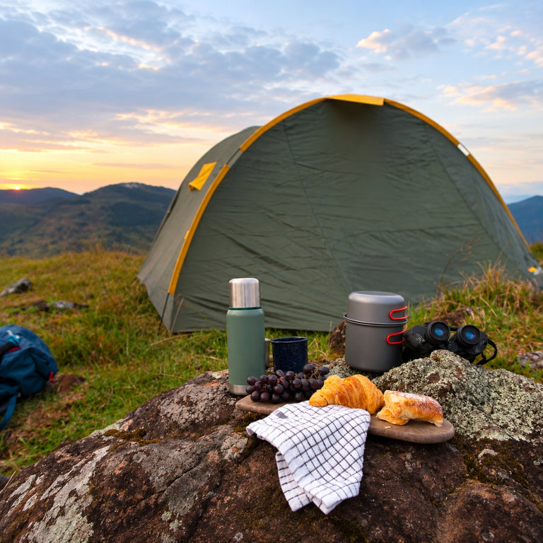 A dome camping tent is pitched on a grassy mountain slope under an overcast sky, with a picnic setup on a rock in the foreground including a thermos, metal pot, bread, grapes, and a checkered towel.