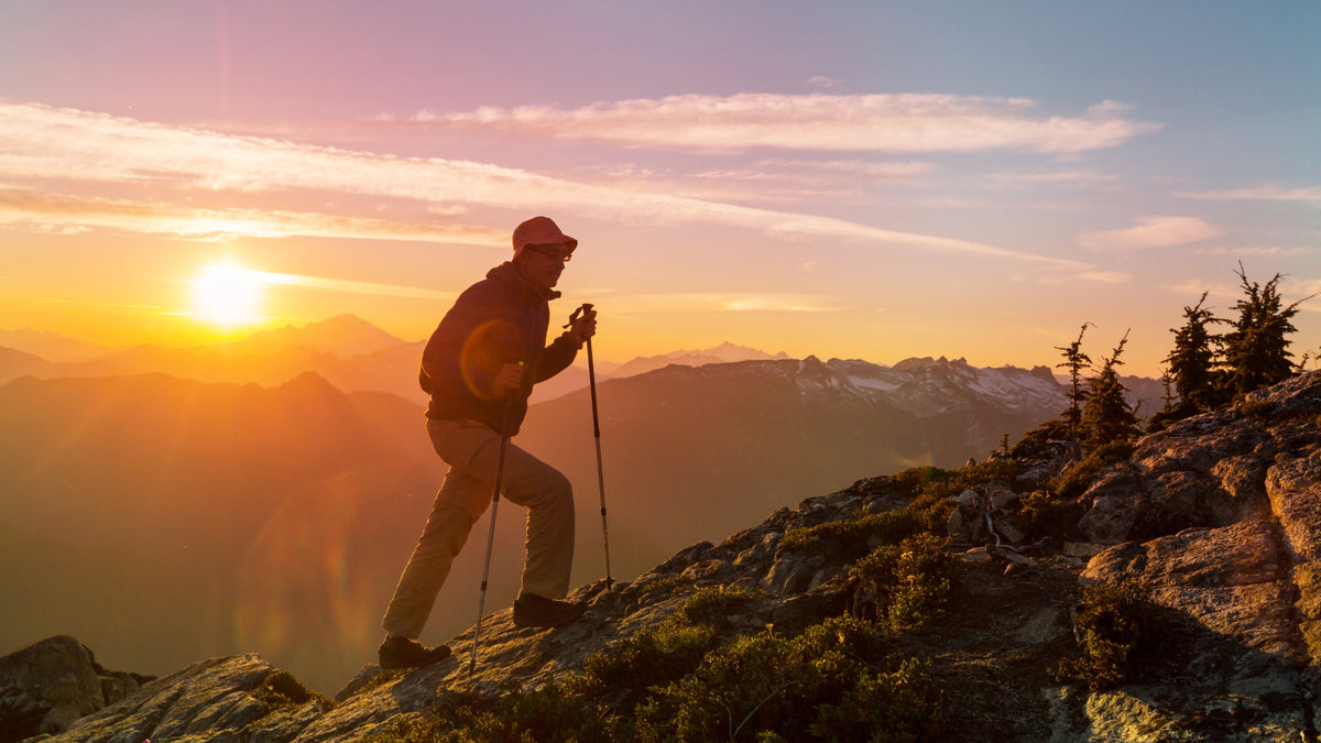 A silhouetted hiker with trekking poles stands on a rocky mountain ridge at sunset, with layered mountain ranges and a glowing sun in the background.